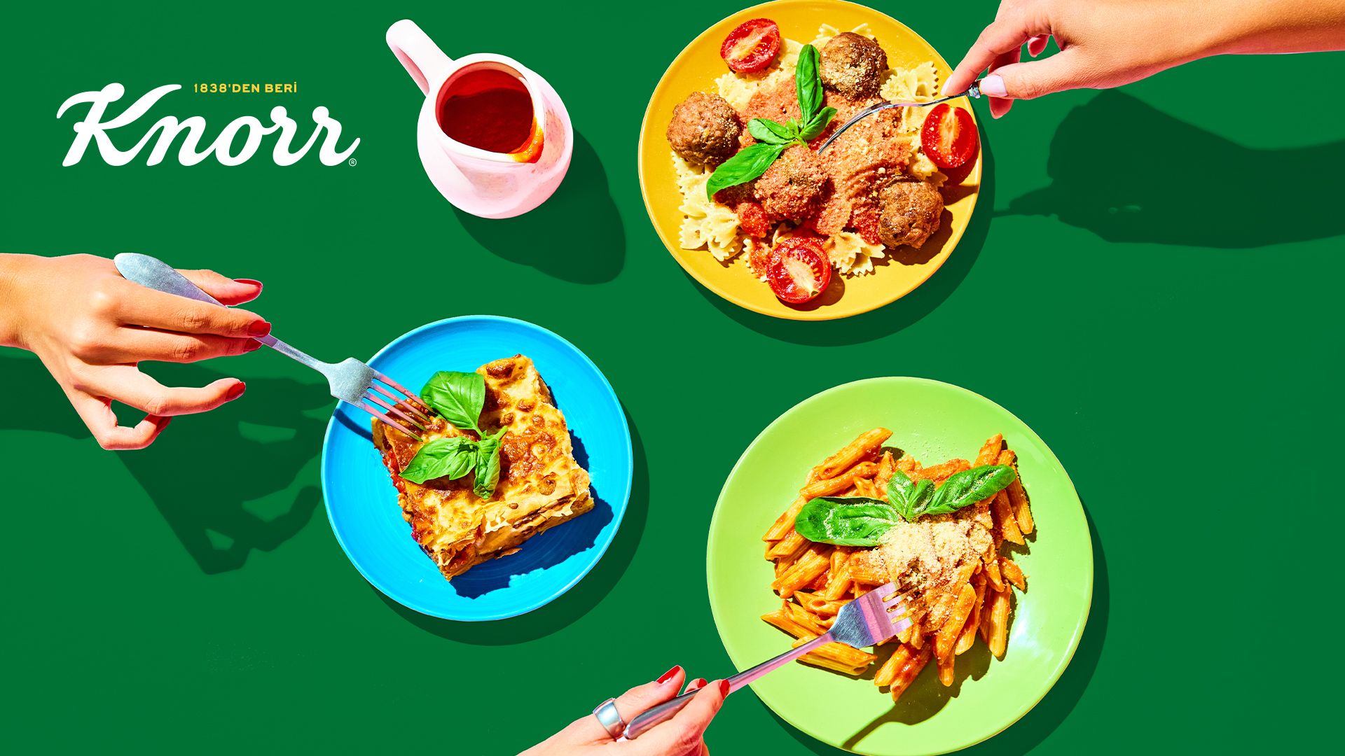 Masadaki tabaklara çatalla uzanan üç elin kuşbakışı görüntüsü.