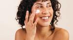 Kısa, koyu renkli ve kıvırcık saçlı bir kadın, parmaklarıyla yanağına az miktarda beyaz yüz kremi uygularken gülümsüyor